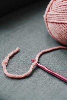 Close-up of a pink crochet hook and yarn ball on fabric, ideal for crafting themes.